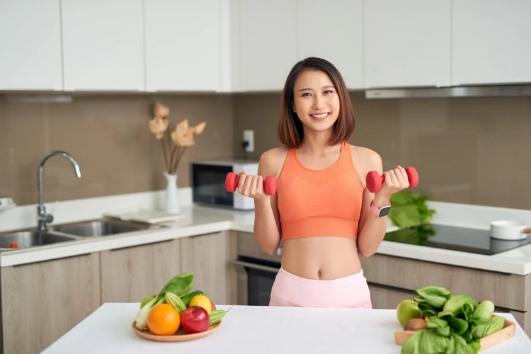 Fit and healthy looking young lady lifting small weights with fruits and vegetables near her.