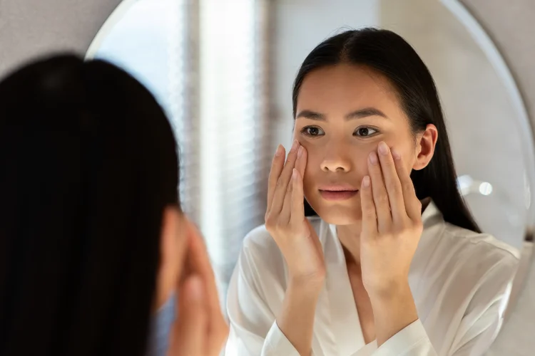 Young Asian woman touching her cheeks in the mirror with even, bright complexion.